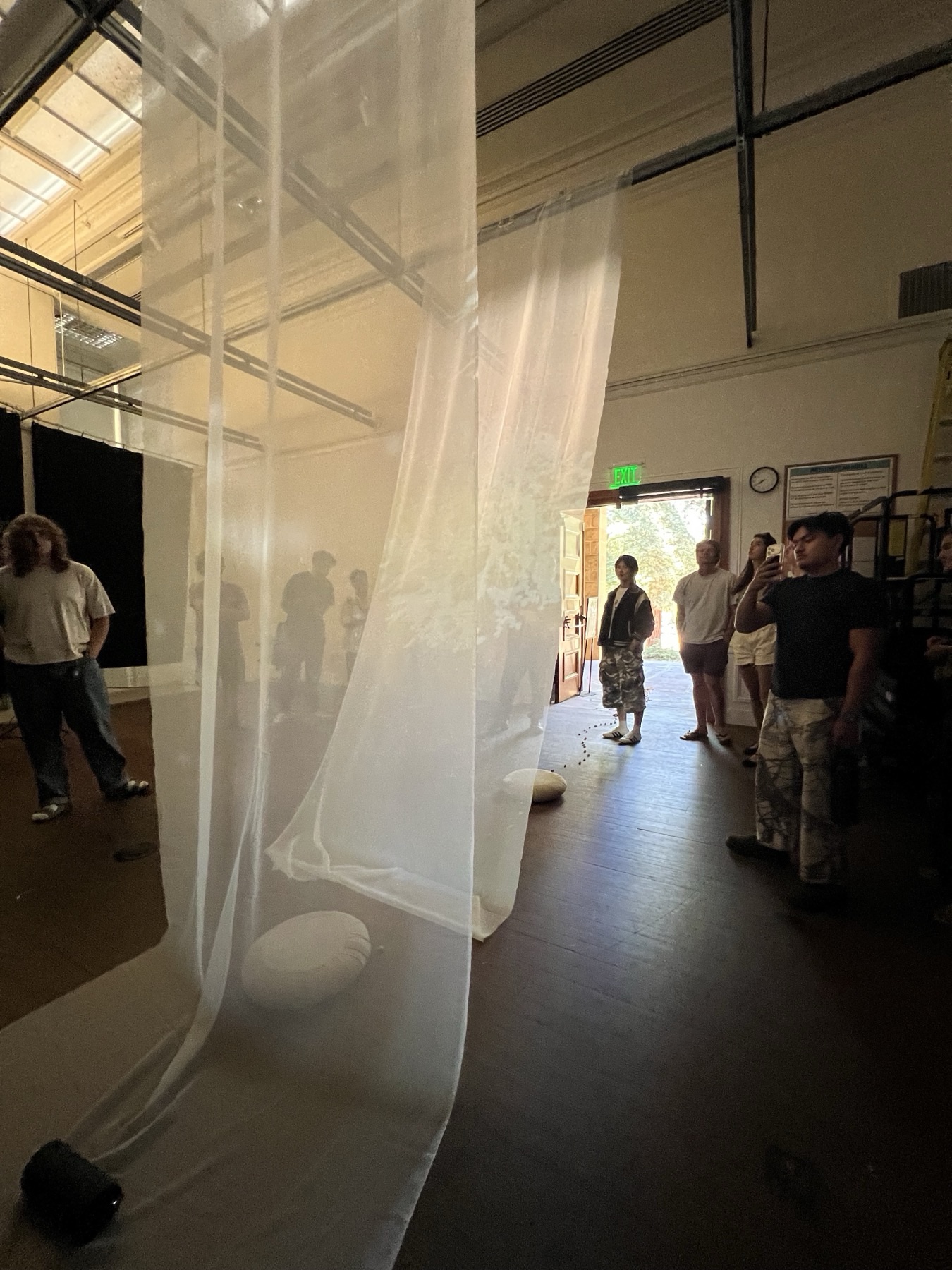 Layers of translucent white fabric hang from the ceiling of a hall, with round cushions on the floor and visitors standing beyond the fabric near an open doorway.